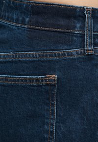 Κοντινή λήψη σκούρων μπλε τζιν τζιν με πίσω τσέπη, θηλιά ζώνης και πορτοκαλί ραφή λεπτομέρειας.