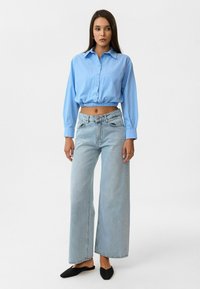 Lichtblauwe cropped shirt met knopen, wijdvallende lichtblauwe jeans en zwarte instap schoenen, staand voor een neutrale achtergrond.