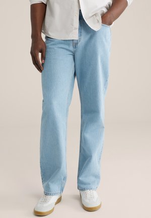 Személy világoskék egyenes szárú farmernadrágban, fehér, gumitalpú tornacipőben és fehér gombos ingben, egyik kezét a zsebében tartva.