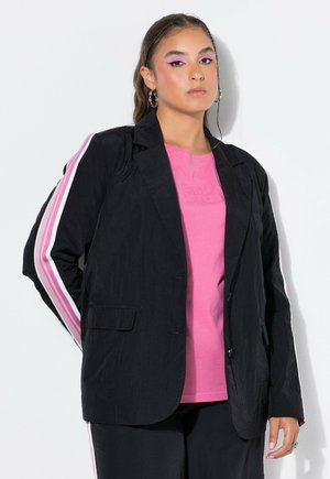 Junge Frau trägt einen schwarzen Blazer mit pinken und weißen Streifen an den Ärmeln über einem pinkfarbenen Hemd und steht vor einem weißen Hintergrund.