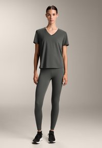 Tummanvihreä urheilupaita, jossa on V-pääntie, yhdistettyinä samanvärisiin tiukkoihinlegginsseihin. Tyylitelty mustilla urheilukengillä. Sileä pinta, rento muotoilu.