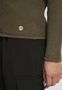 Pull en tricot texturé vert avec un badge en bois rond, porté sur un pantalon noir ajusté, montrant les détails des manches et des coutures latérales.