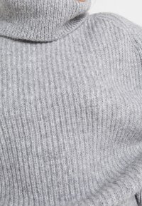 Harmaa pooloneule, valmistettu ribbineuloksesta, jossa korkea kaulus ja pehmeä koostumus. Näkyvät saumat ja tiukka istuvuus.