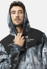 Zwart en blauw tie-dye hoodie met een grafische print en verstelbare trekkoorden, voorzien van gestructureerde mouwen en contrasterende accenten.