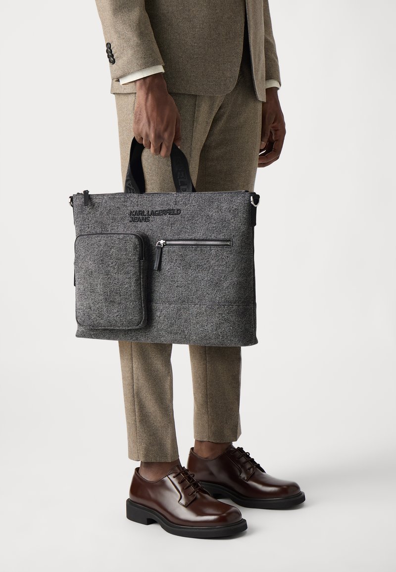 Siva laptop torba s črnim zadrgo, sprednjim žepom in ročaji. Vtisnjen logotip "Karl Lagerfeld Jeans" na sprednji strani.
