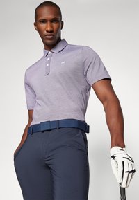 Krátký rukáv fialového polo trička s knoflíkovou légou, v kombinaci s navy kalhotami a tkaným opaskem se čtvercovou sponou. Viditelná bílá golfová rukavice a hůl.