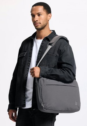 Mand iført sort jakke og hvid skjorte holder grå skuldertaske med polstret rem mod hvid baggrund.