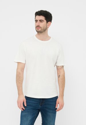 Sötét hajú, szakállas férfi, egyszínű fehér pólót és kék farmert visel, enyhén balra néz fehér háttér előtt.