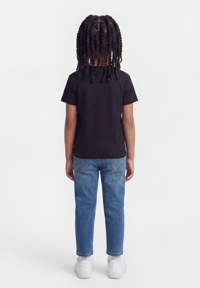 Παιδί με μακριά στριφτά μαλλιά, φορώντας μαύρο μπλουζάκι, μπλε τζιν και λευκά αθλητικά παπούτσια, στέκεται με την πλάτη γυρισμένη σε λευκό φόντο.