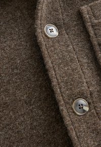 Tissu brun texturé avec une apparence duveteuse, featuring un grand bouton rond en forme de perle avec quatre trous.