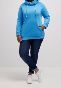 Sudadera azul claro con capucha y cordones ajustables, bolsillos laterales y mangas largas, combinada con vaqueros azul oscuro y zapatillas blancas.