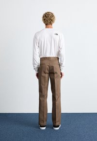 Hnědé kalhoty se střihovými nohavicemi, dvěma zadními kapsami a poutkem na pásek. Skvěle doplněné bílou košilí s dlouhým rukávem, která má logo.