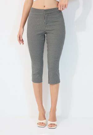 Woman wearing black and white checkered cropped pants and white open-toe heels, standing against a plain white background.