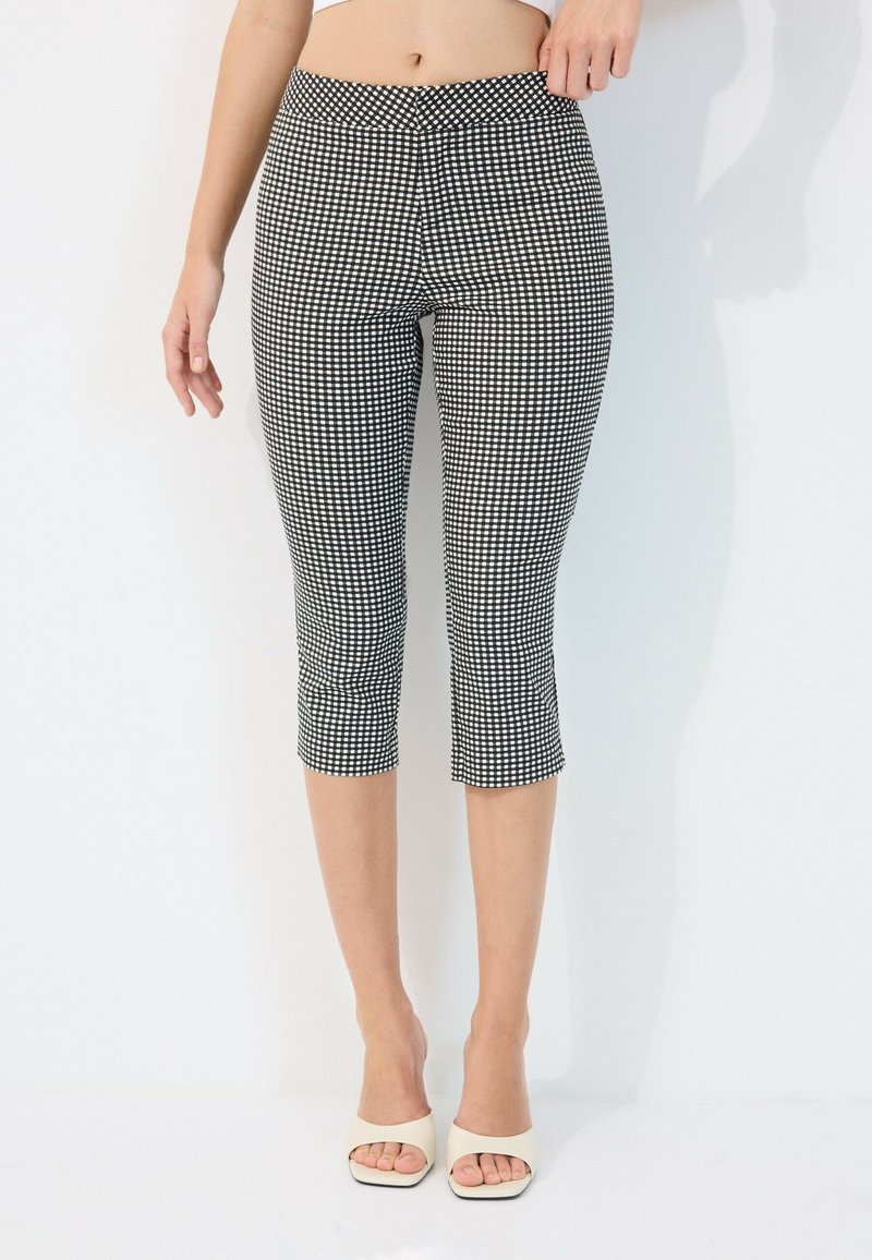 Femme portant un pantalon court à carreaux noir et blanc et des talons blancs ouverts aux orteils, debout sur un fond blanc uni.