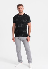 Schwarzes T-Shirt mit kurzen Ärmeln und weißem Sprenkelmuster, kombiniert mit hellgrauen Hosen, getragen mit weißen Sneakers. Einfaches, figurbetontes Design.