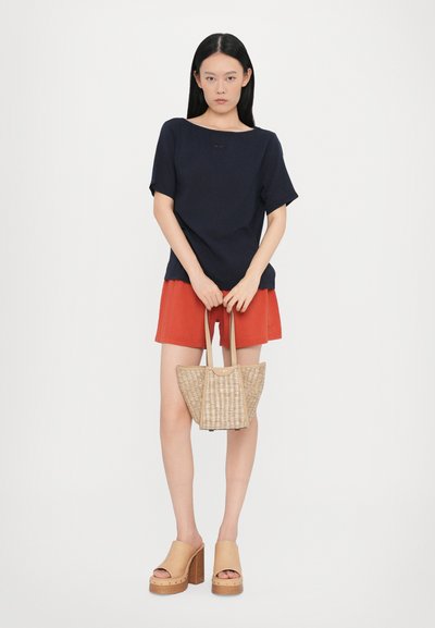 Maglietta blu navy a maniche corte, pantaloni corti rossi, sandali con tacco chunky color beige e borsa di paglia intrecciata. Design semplice, texture liscia, accessori pratici.