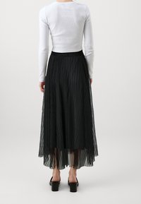 Černá plisovaná sukně s průhledným přehozem, vysokým pasem a vrstvenou strukturou, spárovaná s přiléhavým bílým topem s dlouhým rukávem a černými blokovými podpatky.