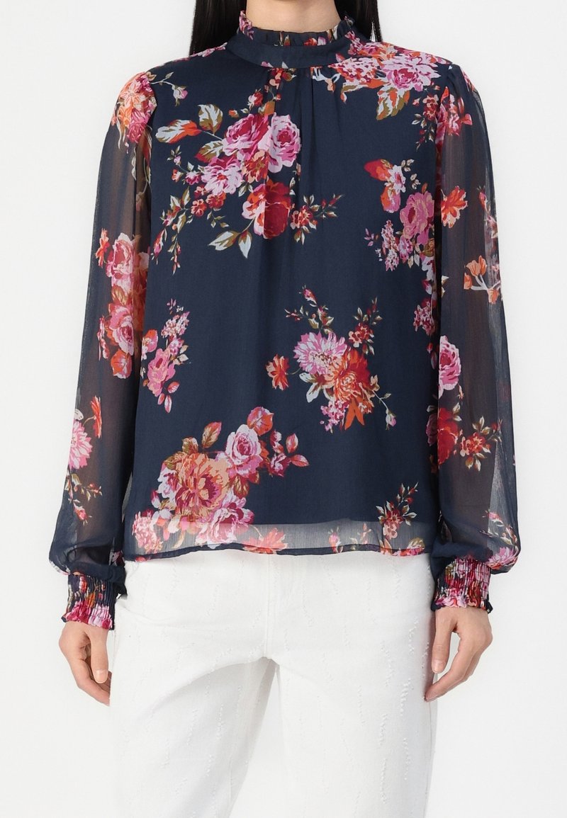 Blouse longue en voile marine à manches longues avec des imprimés floraux roses et rouges, col haut et poignets smockés, associée à un pantalon blanc texturé.