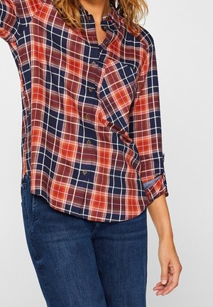 Kvinde iført langærmet ternet skjorte i rød, marineblå og hvid kombineret med blå jeans, med den ene arm løftet og synlig fra talje til nakke.