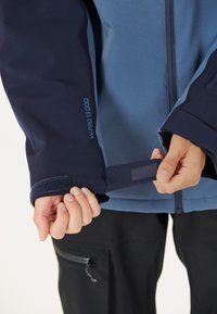 Blau und marineblauer Jacke mit verstellbaren Klettverschlüssen, W-PRO 15000 Branding und einer weichen Textur. Reißverschluss vorne und dunkle Hosen im Hintergrund.