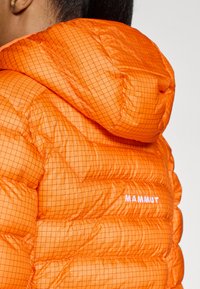 Oranžs pūkains džemperis ar režģa rakstu, ar kapuci un balto "MAMMUT" logotipu sānos. Gluda tekstūra ar siltumizolētām daļām.