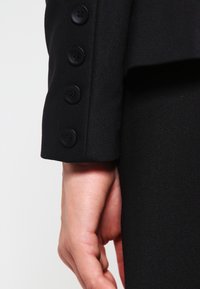 Manica di un blazer nero sartoriale con sei bottoni neri lungo il polsino, texture del tessuto liscia e design strutturato.