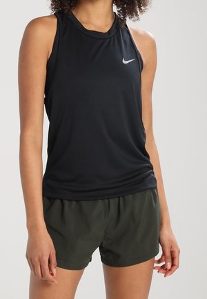 Femme portant un débardeur sportif noir sans manches Nike et un short de course vert foncé, debout devant un fond uni.