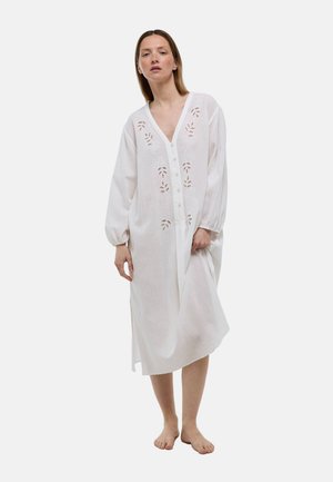 Femme debout pieds nus portant une robe midi blanche à manches longues avec des découpes florales brodées et des boutons sur le devant.