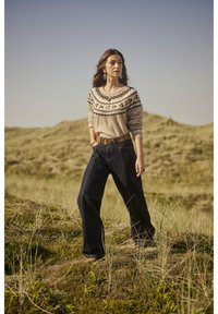 Adīta džemperis ar apaļu kakla izgriezumu, gaiši brūns ar krāsainu rakstu, saskaņots ar brīviem tumši ziliem džinsiem un brūnu jostu; modelis stāv garajā zālē.