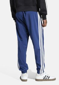 Pantalones deportivos azules con franjas laterales blancas, puños elásticos y una textura suave, combinados con zapatillas blancas con acentos negros.