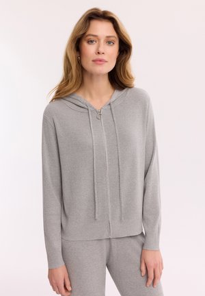 Kobieta o jasnobrązowych włosach, nosząca szarą dzianinową bluzę z kapturem na zamek błyskawiczny i pasujące spodnie, stojąca na jednolitym jasnym tle.