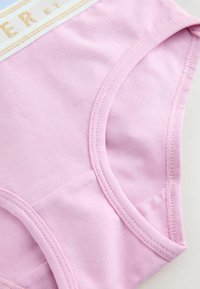 Gros plan sur un sous-vêtement en coton rose clair avec une ceinture élastique ornée d’un texte doré et des coutures renforcées autour des ouvertures des jambes.
