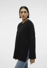 Maglione nero lavorato a maglia con scollatura rotonda, maniche lunghe, spacchi laterali e vestibilità comoda. Indossato con jeans azzurri chiari.