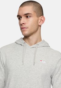 Felpa con cappuccio grigio chiaro realizzata in tessuto misto cotone. Presenta un cappuccio con cordino, dettagli a coste e il logo Fila in bianco e rosso sul petto.