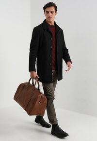 Černý vlněný kabát s knoflíky, burgundský svetr, olivové kalhoty, hnědá kožená taška a černé kotníkové boty. Jednoduchý design, praktické doplňky.
