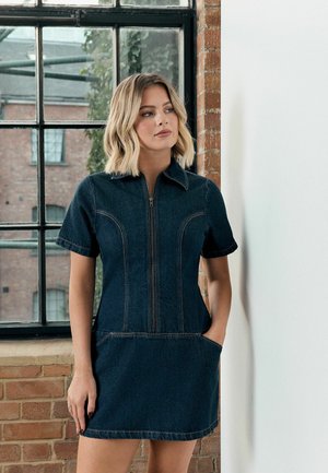 Kvinde med blondt skulderlangt hår iført en kortærmet mørk denimkjole med lynlås foran og lommer, stående ved et vindue og en murstensvæg.