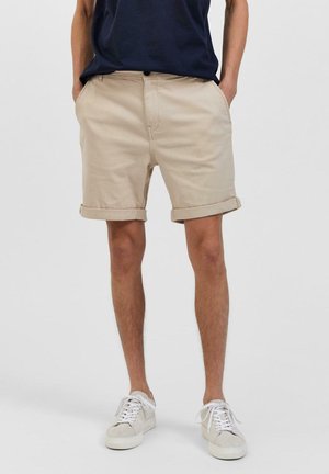 Beżowe bawełniane szorty z podwiniętym dołem, detalem kieszeni z przodu i zapięciem na guzik, w zestawieniu z białymi niskimi trampkami na neutralnym tle.