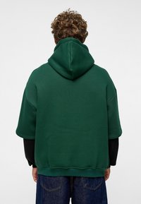Sweat à capuche vert avec une grande capuche, des manches courtes et une coupe décontractée. Fabriqué en tissu doux avec une texture lisse, associé à un jean foncé.