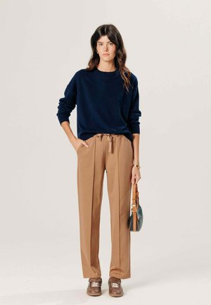 Vrouw draagt een marineblauwe trui, beige wijde broek met trekkoord, bruine sneakers, houdt een kleine blauwe handtas met beige riemen vast, staand tegen een effen achtergrond.