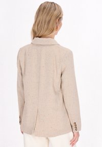 Blazer beige texture con colletto a punte, cuciture visibili e striature colorate; presenta maniche lunghe e polsini con quattro bottoni.