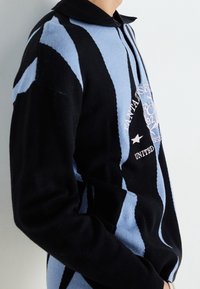 Knitted black and blue pullover with a collared neck, featuring bold vertical stripes and a logo detail on the chest. Soft, textured fabric.