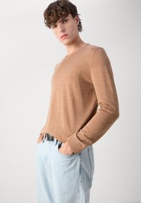 Light brown long-sleeve shirt with a v-neck, made from a soft material, paired with light blue denim jeans and a black belt.