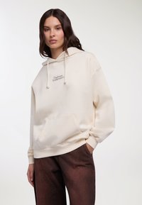 Felpa con cappuccio color crema con tasca frontale, coulisse e dettaglio ricamato "Undercovers Troublemakers". Tessuto morbido e vestibilità rilassata.