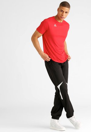 Rotes Sport-T-Shirt mit kurzen Ärmeln und einem kleinen weißen Logo; schwarze Trainingshose mit weißen Akzenten und elasticen Bündchen; weiße Sneakers.