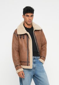Uomo che indossa una giacca di shearling marrone con foderato in pelliccia crema, una camicia nera e jeans blu, in piedi con una mano in tasca su uno sfondo bianco.