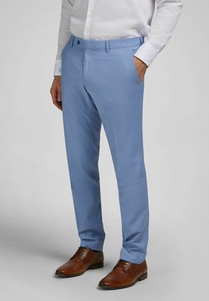 Mann trägt hellblaue Anzughose, weißes Hemd und braune Lederschuhe, steht mit einer Hand in der Tasche.