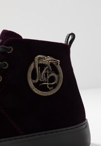 Ciemnofioletowe zamszowe sneakersy za kostkę z czarną podeszwą i brązowym emblematem w kształcie węża z logo "Just Cavalli" na boku.