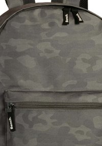 Grijze camouflage rugzak van duurzaam materiaal, met een voorzak met rits, dubbele schouderbanden en logo-accenten. Glad oppervlak en rechthoekige vorm.