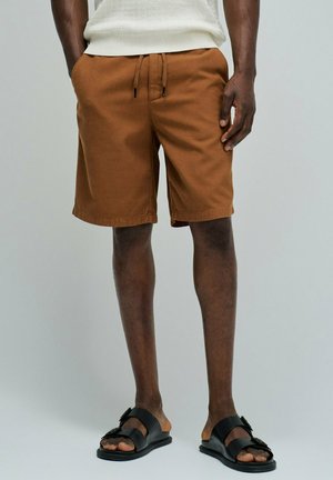 Hombre con pantalones cortos marrones con cordón, camiseta blanca texturizada y sandalias negras con hebilla sobre un fondo claro y liso.