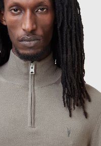 Hellbrauner, mit Reißverschluss versehener Pullover mit Stehkragen und feiner Strickoberfläche. Verfügt über ein kleines Logo auf der Brust und eine glatte Oberfläche.